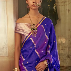 May include: A woman wearing a royal blue sari with gold accents and a matching off-the-shoulder top. The sari features a dotted pattern and gold stripes. She is adorned with gold jewelry, including a necklace, earrings, nose ring, bracelet, and ring.