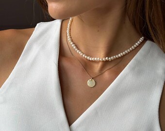 May include: A woman wearing a pearl necklace, a gold pendant necklace, and gold hoop earrings with a pearl drop. The necklaces are layered, with the pearl necklace closer to the neck. The woman is wearing a white vest.