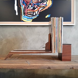 May include: A wooden record stand with a long, narrow shelf and a vertical section for storing records. The stand is made of dark wood and has a natural finish. The stand is holding a stack of vinyl records.