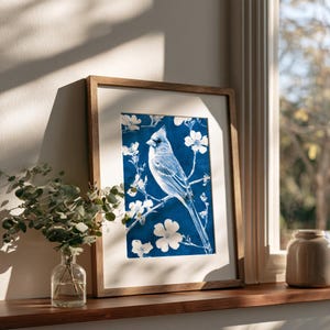 Puede incluir: Una lámina artística enmarcada que muestra un cardenal blanco posado en una rama rodeado de flores blancas sobre un fondo azul intenso. La obra está dentro de un marco de madera sencillo con un paspartú blanco, mostrando un diseño contrastado.
