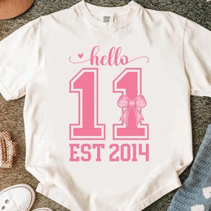 May include: White t-shirt with a pink design. The design includes the word "hello", the number "11", a bow, and the text "EST 2014". The shirt is made of a soft material and has a crew neck.