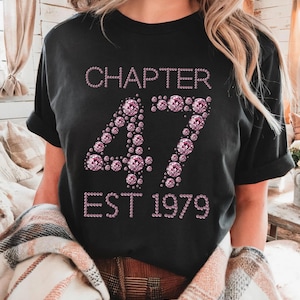 May include: Black t-shirt with the text "CHAPTER 47 EST 1979" in pink rhinestones. The number 47 is prominently displayed in a sparkling, gem-like font. The shirt has short sleeves and a relaxed fit.