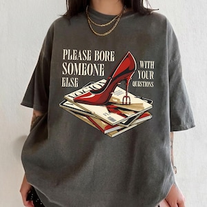 May include: Dark grey t-shirt featuring a graphic of a red high heel shoe atop a stack of magazines. The text on the shirt reads "PLEASE BORE SOMEONE ELSE WITH YOUR QUESTIONS".