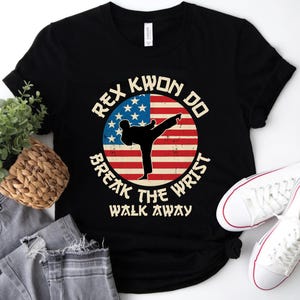 May include: Black t-shirt featuring a graphic of a martial artist kicking, set against a red, white, and blue American flag design. The text reads "REX KWON DO BREAK THE WRIST WALK AWAY".