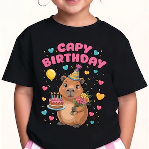 May include: Black t-shirt with a colorful graphic featuring a capybara wearing a party hat and holding a cake and flowers. The text "CAPY BIRTHDAY" is above the capybara, with hearts and confetti scattered around.