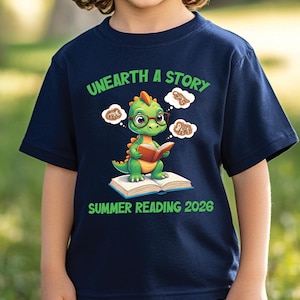 May include: Navy blue t-shirt featuring a cartoon dinosaur reading a book, wearing glasses, and surrounded by thought bubbles. The shirt reads "UNEARTH A STORY" and "SUMMER READING 2026" in green text.