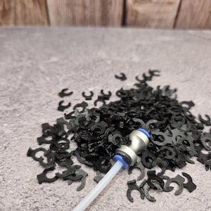 May include: A large pile of small black C-shaped plastic clips scattered across a surface. A single silver and blue pneumatic tube fitting with a short length of white tubing is resting on top of the collection of black plastic fasteners.
