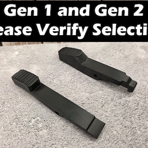 May include: Two black, elongated, matte-finished components with textured, ribbed grip sections at one end. The text 'Gen 1 and Gen 2 Please Verify Selection' is displayed in white letters on a black background above the two items.