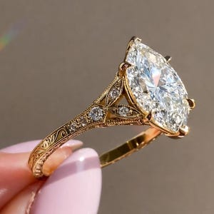 Puede incluir: Un anillo de compromiso dorado con un gran diamante en forma de pera. La banda está adornada con diamantes más pequeños y grabados detallados. El anillo tiene un diseño vintage, que resalta el brillo del diamante central. Una joya elegante.