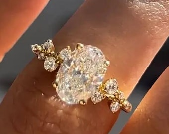 May include: A gold ring featuring a large oval-cut center stone flanked by smaller round-cut stones on each side. The band is crafted from polished yellow gold, and the setting includes a cluster of accent stones arranged in a leaf-like pattern.