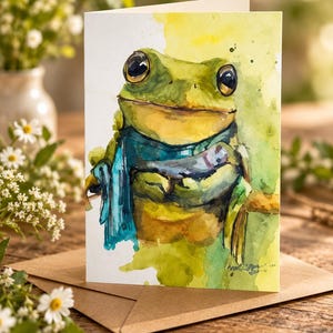 May include: A greeting card featuring a watercolor illustration of a green frog with large dark eyes wearing a blue scarf. The frog is depicted against a soft yellow and green wash background, with visible brushstrokes and paint splatters.