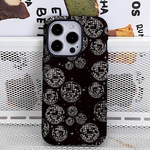 May include: A black phone case featuring a pattern of disco balls in shades of gray and white. The case has a dark background with small star accents. The phone's camera lenses are visible at the top.