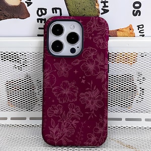May include: A burgundy phone case with a floral design. The case features a pattern of outlined flowers and stars. The phone case has a black border around the camera and a black border around the edge of the phone.