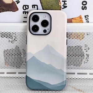 Puede incluir: Funda de teléfono con un diseño de paisaje montañoso. La funda tiene un fondo blanco con un degradado de montañas azules. El área de la cámara es negra con tres lentes. La funda está hecha de un material liso y brillante.