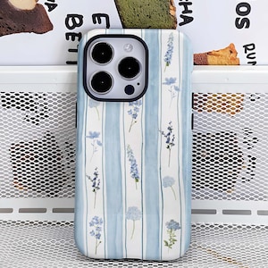 Peut inclure: Une coque de téléphone avec un motif floral. L'étui présente un motif de rayures verticales bleu clair avec de délicates illustrations de fleurs bleues. L'étui de téléphone a un bord noir autour de la caméra et un fond blanc.