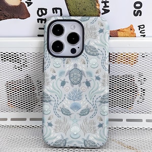 May include: A phone case with a sea turtle pattern. The case features a light blue background with a repeating design of sea turtles, seashells, and coral in shades of blue, gray, and white. The case has a black border around the camera.