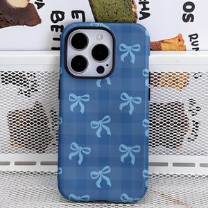 Pode incluir: Uma capa de telefone azul com um padrão xadrez e laços azuis claros. A capa é projetada para um smartphone e possui um recorte para a câmera. O design é de estilo fofo e feminino.