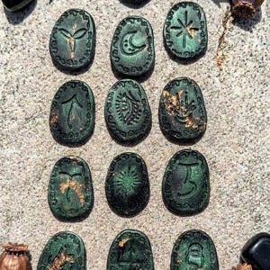 Könnte beinhalten: Ein Set aus dreizehn ovalen Steinen in dunkelgrüner Ausführung mit metallischen Goldakzenten. Jeder Stein weist ein einzigartiges eingraviertes Symbol auf seiner Oberfläche auf. Die Sammlung ist gitterförmig angeordnet und von kupferfarbenen Mohnkapseln umgeben.