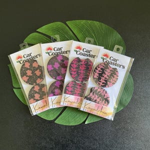 May include: Four packs of car coasters with floral designs in pink, purple, and black. Each coaster is packaged in a clear plastic sleeve with the words "Car Coasters" and "Kawaile Collective" printed on the packaging.