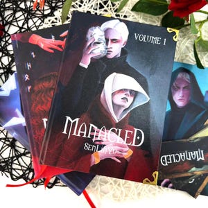 Op de afbeelding: Een stapel fantasieromans met opvallende omslagillustraties. Het hoofdboek toont een donkere illustratie van twee figuren, een met een masker, met de titel "Manacled" en "Volume 1". De covers hebben gouden hoekbeschermers en rode lint-bladwijzers.
