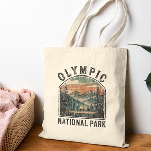 Puede incluir: Bolso tote de color crema con un gráfico desgastado de un paisaje montañoso y el texto "OLYMPIC NATIONAL PARK" en negro. El bolso tiene dos asas y está hecho de una tela resistente, perfecto para llevar objetos.