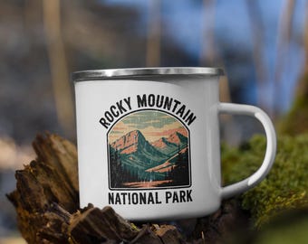 May include: White enamel mug with a silver rim and handle. The mug features a graphic of a mountain landscape with the text "Rocky Mountain National Park" in black and green. The mug is sitting on a piece of wood.