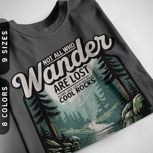 Op de afbeelding: Donkergrijs t-shirt met een thema-afbeelding van de natuur. Het ontwerp bevat de woorden "Not All Who Wander Are Lost" boven een schilderachtige illustratie van een bos en een pad. De tekst "Some Are Looking For Cool Rocks" is ook opgenomen.
