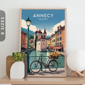 May include: A framed art print with a vintage travel poster style illustration of Annecy, France. The artwork shows a canal scene with buildings, a bicycle with flowers, and the text "Annecy France".