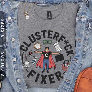 Könnte beinhalten: Graues T-Shirt mit einem Grafikdesign, das "CLUSTERF*CK FIXER" in einem kreisförmigen Design zeigt. Das Design zeigt einen Cartoon-Superhelden mit rotem Umhang, umgeben von Symbolen wie Geld, einer Uhr und einer Aktentasche.