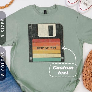 May include: A sage green t-shirt featuring a retro design of a floppy disk with the text "BEST OF 1979" in a colorful striped pattern. The shirt has a vintage aesthetic, suggesting a nostalgic theme.