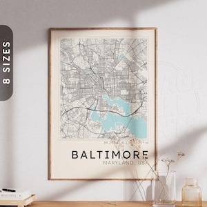May include: A framed wall art print featuring a detailed street map of Baltimore, Maryland, USA. The map displays black road networks on a cream background with light blue water areas. Text includes the city name, state, and coordinates 39.2904 N | 76.6125 W.