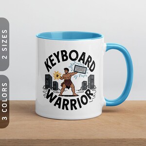 May include: White ceramic mug with a blue interior and handle. The mug features the words "KEYBOARD WARRIOR" in black, with a cartoon illustration of a muscular figure holding a keyboard. The mug has a light brown wooden surface.