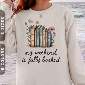 May include: Cream-coloured sweatshirt with a graphic of colourful books and flowers. The text "my weekend is fully booked" is printed below the illustration. Available in 8 sizes and 6 colours.