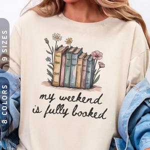 May include: Cream-coloured long-sleeved sweatshirt with a graphic of books and flowers, and the text "my weekend is fully booked". The design features a stack of books in pastel colours and floral elements.