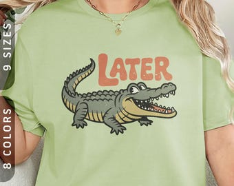 May include: Light green t-shirt featuring a cartoon alligator with the word "LATER" in orange letters. The alligator is gray and yellow with a smiling expression. The shirt also has text that says "9 sizes" and "8 colors".