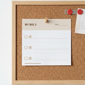 Könnte beinhalten: Eine quadratische Aufgabenliste aus Papier mit der Überschrift MY BIG 3, drei nummerierten Kästchen und Schreibzeilen, befestigt an einer rechteckigen Pinnwand aus Kork mit hellem Holzrahmen. Ein kleiner Kalender mit roten Magneten ist ebenfalls angebracht.