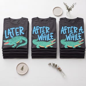 Puede incluir: Tres pilas de camisetas negras con gráficos de caimanes. Cada camiseta tiene una frase diferente en azul: "LATER", "AFTER WHILE" y "AFTER A WHILE". Los caimanes son verdes con bocas rojas.