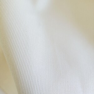 May include: A close-up view of off-white fabric featuring a distinct diagonal twill weave pattern. The material has a dense, uniform texture with visible parallel ridges running across the surface, creating a consistent and durable appearance.