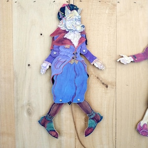May include: A flat, articulated paper doll figure depicting a white wolf with a monocle, wearing a purple jacket with a red collar, a blue skirt, and brown leggings with teal and red shoes. The figure features metal rivets at the joints.