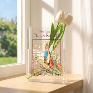 May include: A clear acrylic vase featuring an illustration from "The Tale of Peter Rabbit." The vase holds white tulips. The illustration depicts Peter Rabbit in a blue jacket and red scarf. The vase is on a wooden surface near a window.