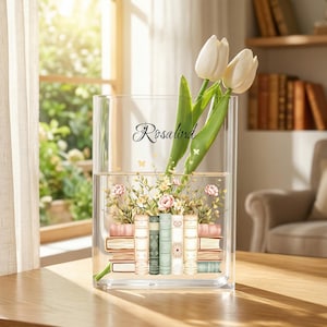 May include: A clear, rectangular vase with the name "Rosalind" in script. The vase holds white tulips and a floral arrangement of pink roses and greenery. The vase is decorated with a stack of books.