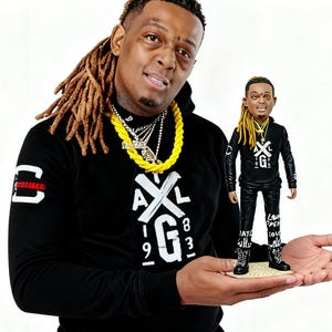 May include: A person holding a miniature figurine. The person is wearing a black hoodie with white text and a yellow necklace. The figurine is dressed in a similar outfit, with black pants and boots, and has the same hairstyle as the person.