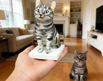 May include: A detailed figurine of a gray tabby cat with green eyes, perched on a white square base. The cat has black stripes and is in a seated position. Another cat of similar appearance is in the background.