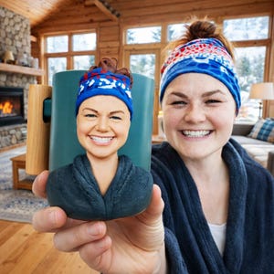 Può includere: Una tazza in ceramica verde acqua con manico in legno. La tazza presenta una somiglianza scolpita in 3D di una persona che indossa una fascia a fantasia blu e rossa e un accappatoio blu scuro. La persona nell'immagine indossa anche una fascia abbinata.