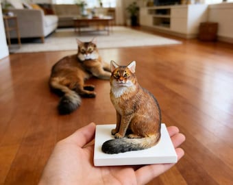 May include: A detailed, brown and black cat figurine sits on a white square base, held in a person's hand. Another cat rests in the background. The figurine has a realistic appearance, with detailed fur and facial features. The cat's tail is black.