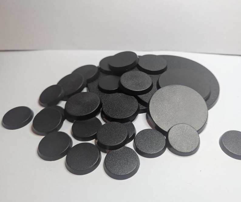 Puede incluir: Un conjunto de bases circulares negras de varios tama&ntilde;os. Las bases tienen un acabado mate y est&aacute;n dispuestas en una pila, con algunas dispersas. Las bases son probablemente para miniaturas o modelos.