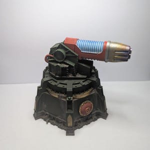 May include: A detailed model of a futuristic cannon. The cannon features a red and green body with a blue and silver barrel. The base is dark green with gold accents and a red circular emblem.