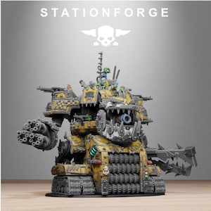 May include: A StationForge miniature tank featuring a yellow and black checkered paint scheme with a large, toothy jaw front. The vehicle is equipped with a multi-barreled cannon, a spiked claw arm, and several small green figures positioned on top.
