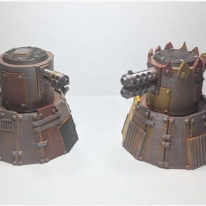 May include: Two miniature, weathered, brown and rust-colored tank-like structures with gun barrels. One has a single barrel, the other has multiple barrels. The bases are a mix of brown and rust-colored panels.