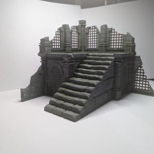 Op de afbeelding: Een grijs miniatuurmodel van een verwoeste stenen structuur, met een trap die leidt naar een platform met gewelfde openingen en een afbrokkelende gevel. Het model heeft een verweerde uitstraling, wat duidt op ouderdom en verval.